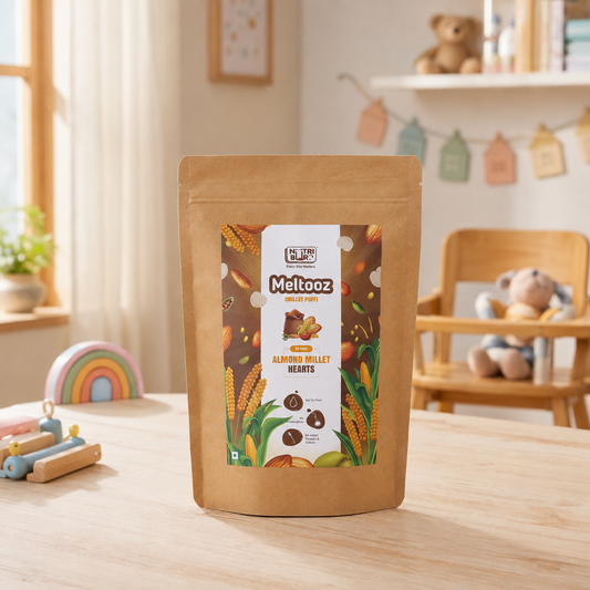 A stand-up kraft paper pouch of Meltooz Almond Butter Hearts chocolate mix, set on a light wooden table in a warm, child-friendly room with toys and a high chair in the background.